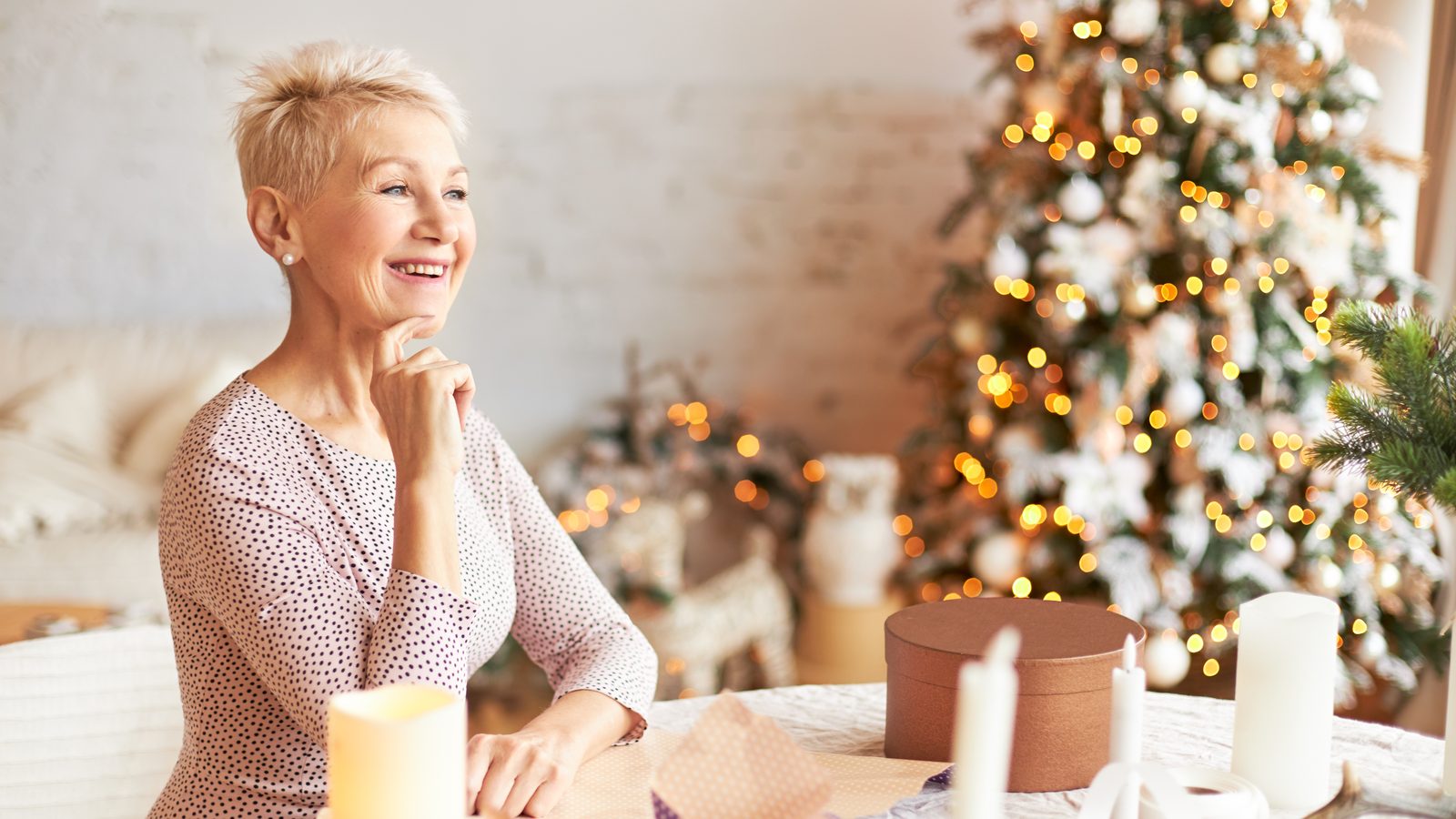 Eine Frau mit kurzen Haaren sitzt laechelnd am Tisch mit Geschenkpapier und Kerzen, dahinter ein beleuchteter Weihnachtsbaum