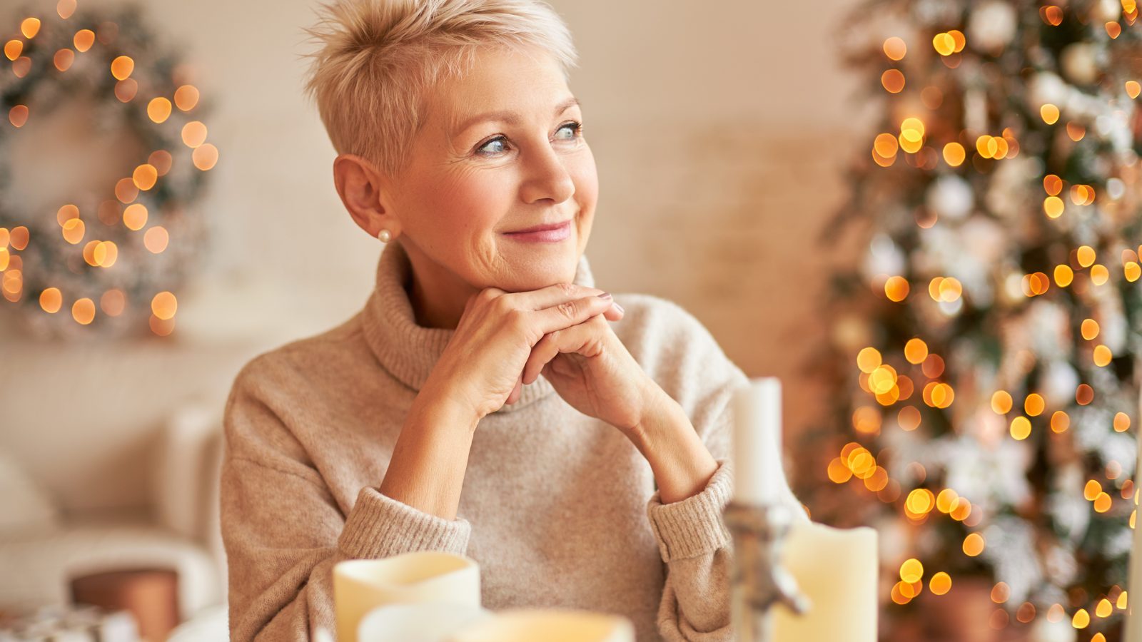 Eine Frau im Pullover sitzt an einem Tisch mit Kerzen und schaut laechelnd zur Seite, im Hintergrund ein Weihnachtsbaum