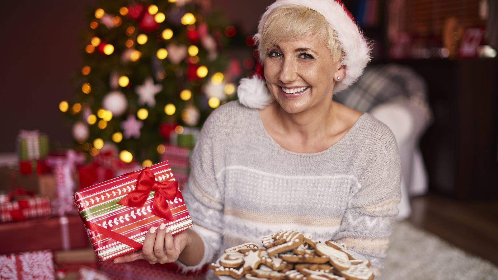 Eine Frau mit Weihnachtsmuetze haelt ein rot eingepacktes Geschenk und einen Teller mit Lebkuchen