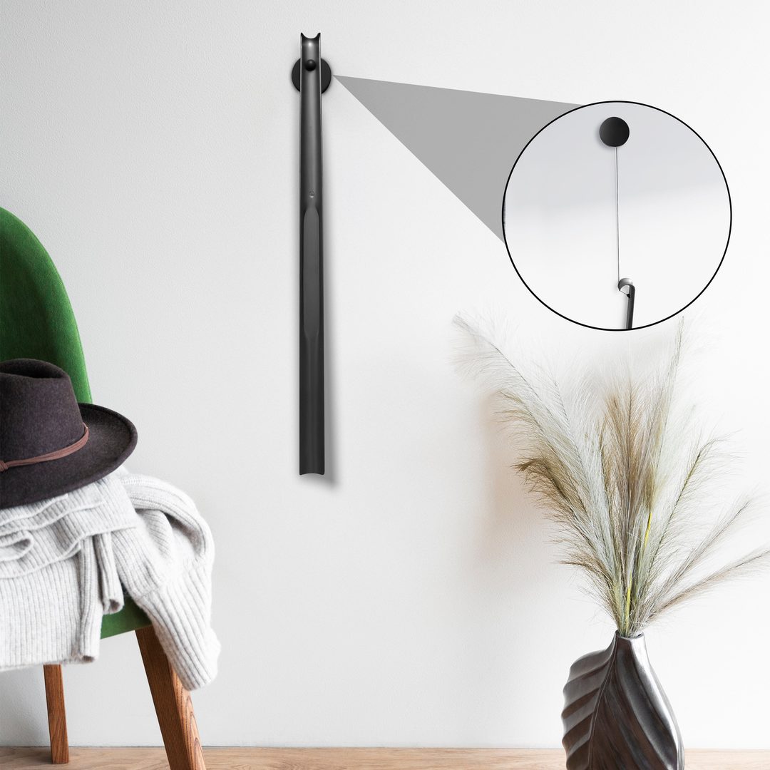 Schuhloeffel haengt eingerollt an einer hellen Wand neben einem Sessel mit Hut, rechts eine Vase mit Graesern