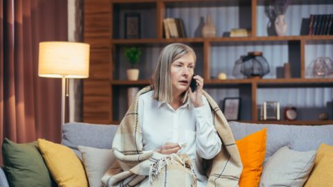 Aeltere Frau sitzt abends in eine Decke gehuellt auf dem Sofa und telefoniert mit besorgtem Blick