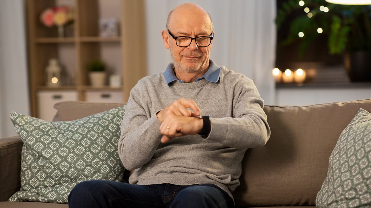 Aelterer Mann mit Brille sitzt auf dem Sofa und sieht auf die Armbanduhr an seinem Handgelenk