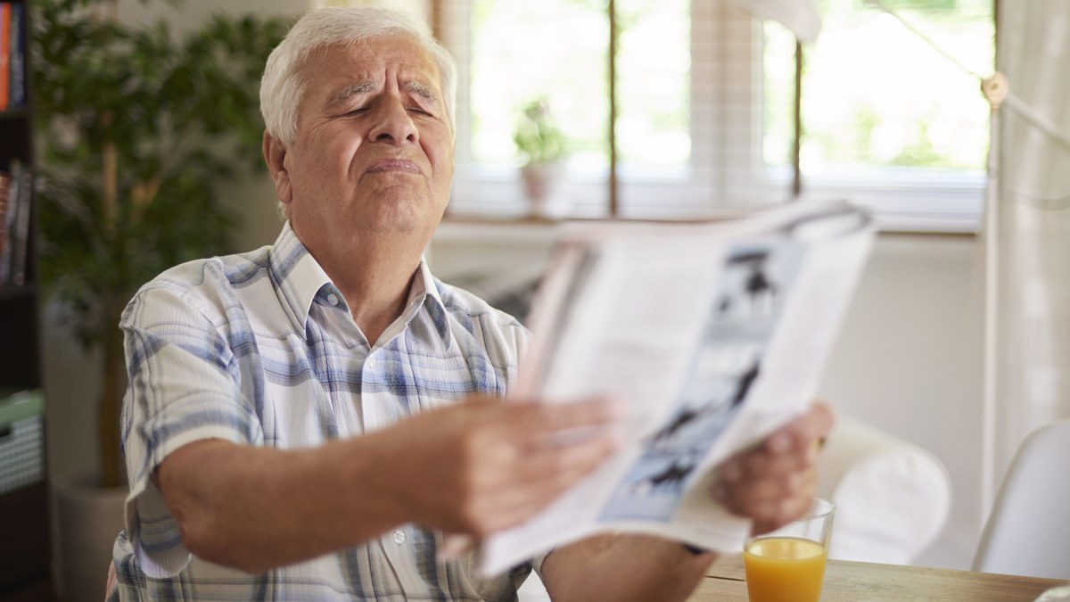 Aelterer Mann haelt die Zeitung mit ausgestreckten Armen weit weg und kneift die Augen zusammen