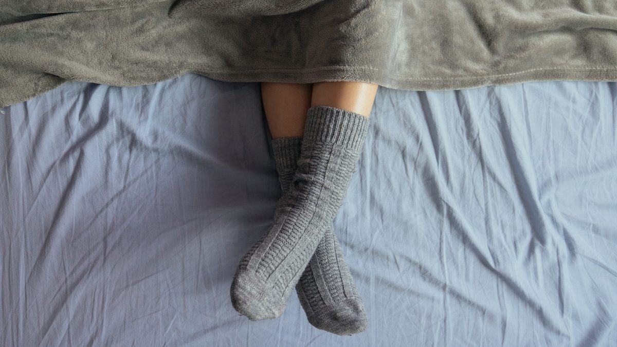 Zwei Fuesse in grauen Strick-Wollsocken schauen unter einer grauen Decke auf einem hellblauen Bettlaken hervor