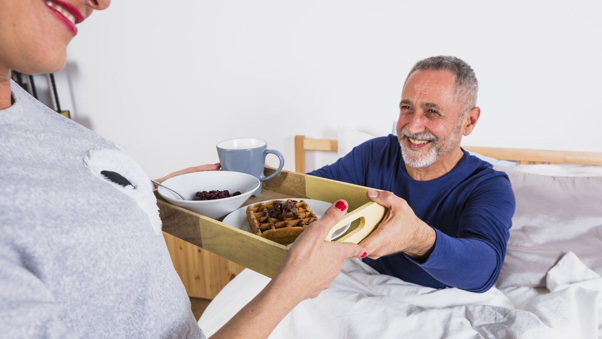 Aelterer Mann sitzt im Bett und nimmt ein Fruehstueckstablett mit Tasse und Waffel von seiner Frau entgegen