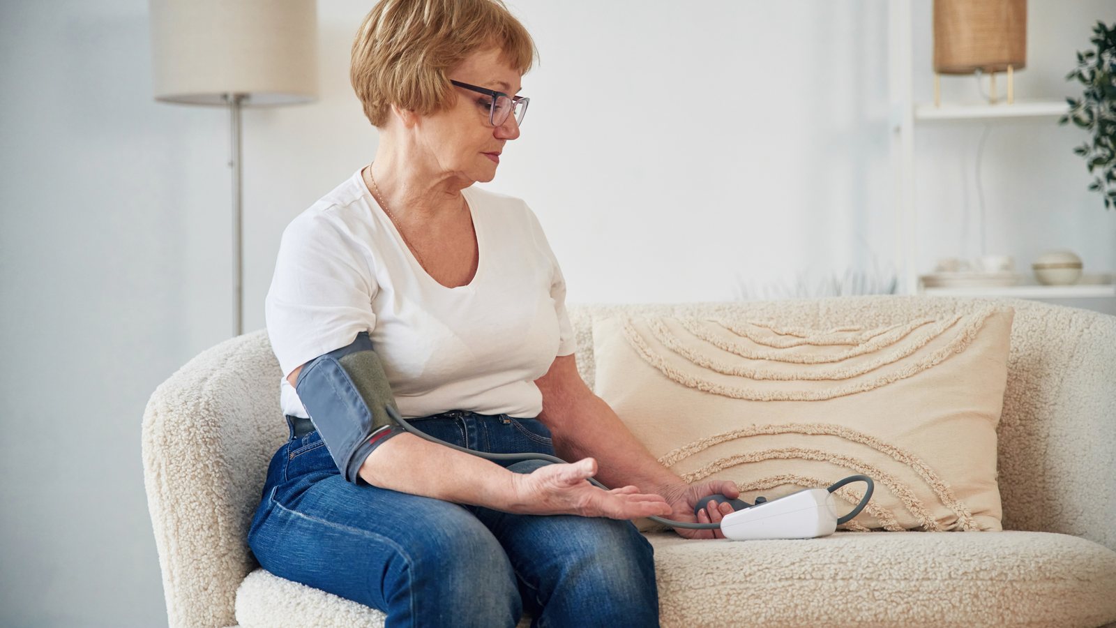 Seniorin sitzt auf dem Sofa und misst ihren Blutdruck mit einem Oberarm-Blutdruckmessgeraet