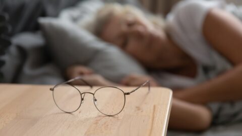 Brille liegt auf einem Holz-Nachttisch, im Hintergrund schlaeft eine aeltere Frau im Bett