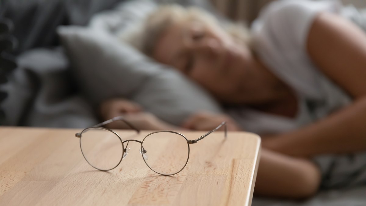 Brille liegt auf einem Holz-Nachttisch, im Hintergrund schlaeft eine aeltere Frau im Bett