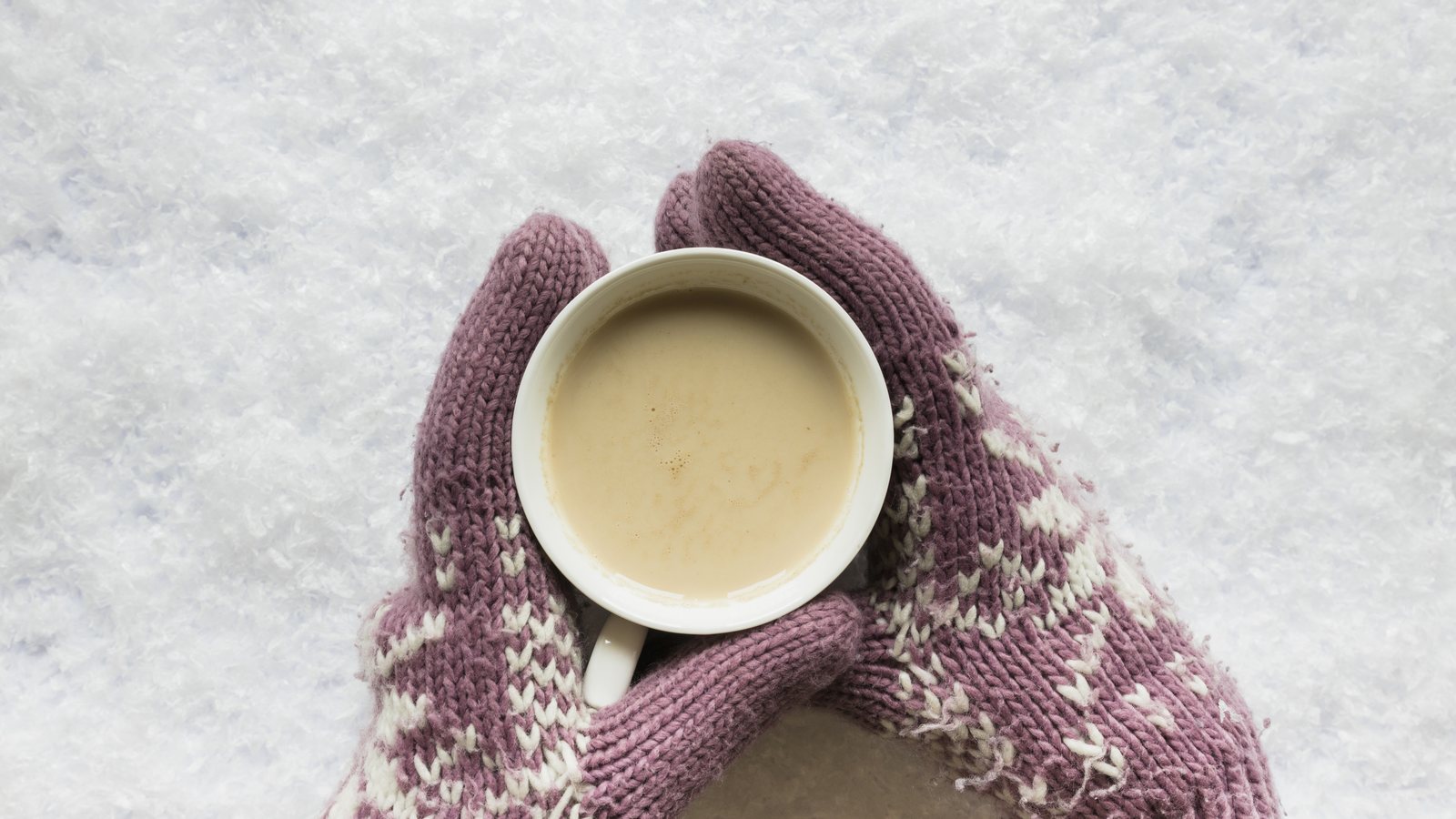 Haende in gestrickten Faeustlingen mit Norwegermuster halten eine Tasse, im Hintergrund Schnee