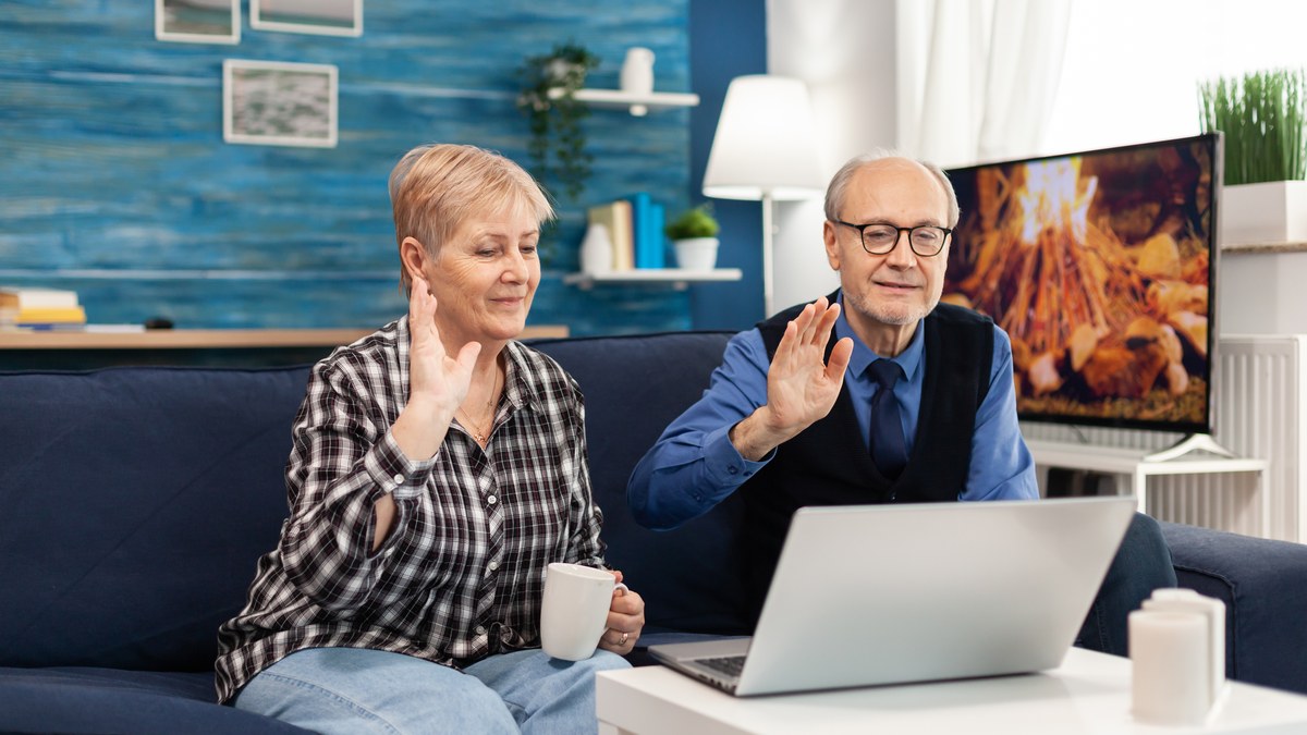 Aelteres Ehepaar auf dem Sofa winkt bei einem Videoanruf in den Laptop