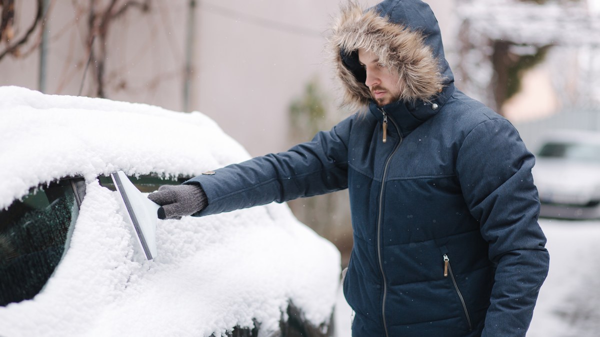 Mann in Winterjacke kratzt die vereiste Seitenscheibe eines verschneiten Autos frei