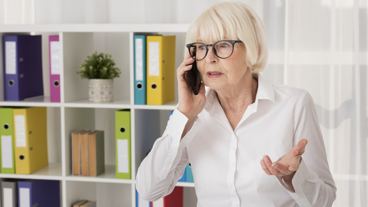 Seniorin mit Brille telefoniert und hebt fragend die Hand, im Hintergrund Aktenordner