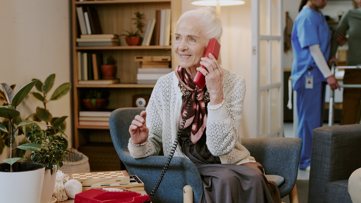 Aeltere Frau sitzt im Sessel und telefoniert laechelnd mit einem roten Festnetztelefon