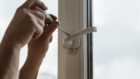 Zwei Haende schrauben mit einem Schraubendreher einen abschliessbaren Griff an ein weisses Kunststofffenster