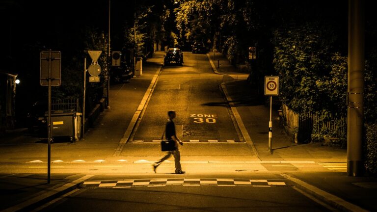 Fussgaenger in dunkler Kleidung ueberquert abends eine nur von Laternen beleuchtete Strasse