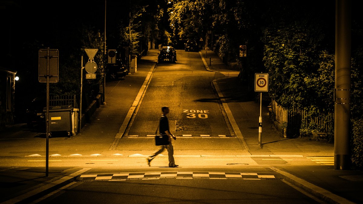 Fussgaenger in dunkler Kleidung ueberquert abends eine nur von Laternen beleuchtete Strasse