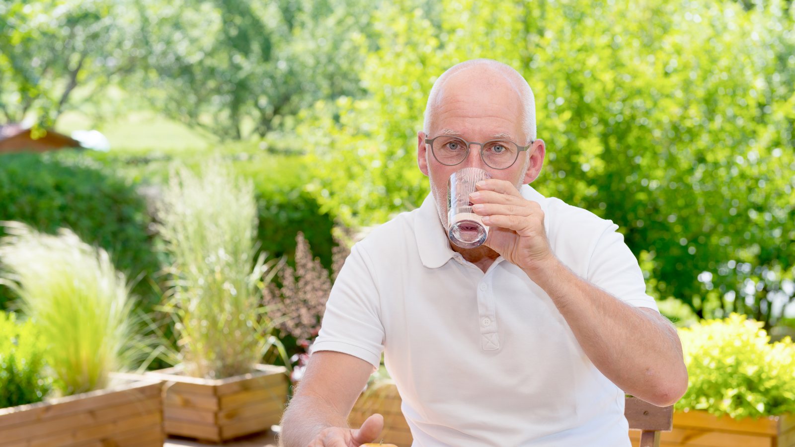 Aelterer Mann mit Brille sitzt im Sommer auf der Gartenterrasse und trinkt ein Glas Wasser