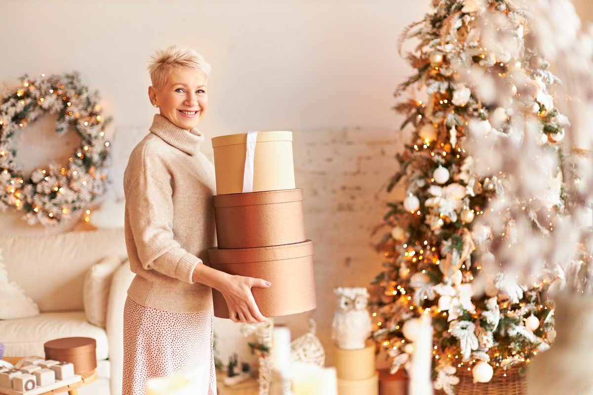 Frau traegt drei Geschenkschachteln durch ein weihnachtlich geschmuecktes Wohnzimmer