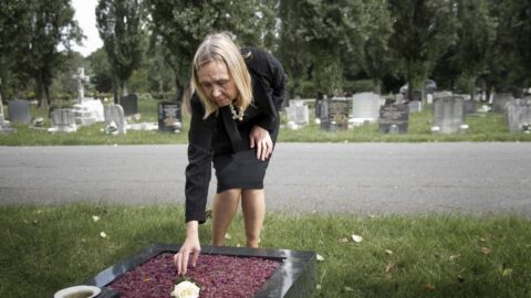 Aeltere Frau beugt sich auf einem Friedhof ueber ein Grab und legt eine Bluete ab