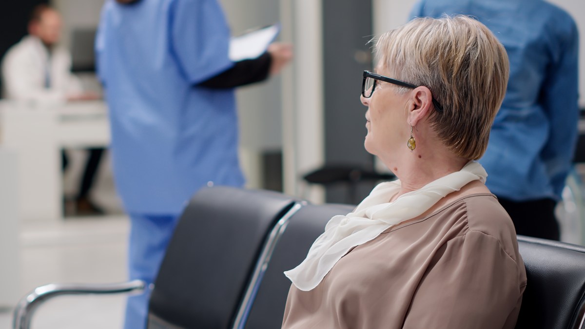 Aeltere Frau mit Brille sitzt im Wartebereich einer Arztpraxis, im Hintergrund Praxispersonal