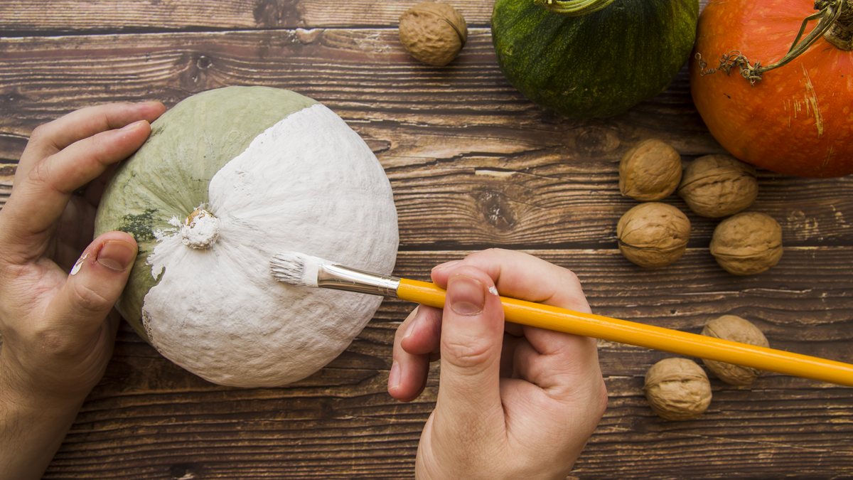 Haende bemalen einen kleinen Kuerbis mit Pinsel und weisser Farbe auf einem Holztisch