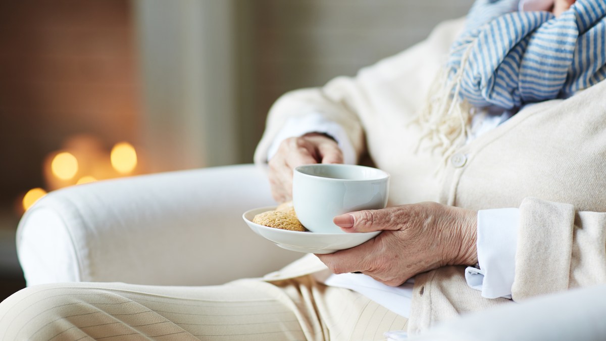 Aeltere Frau in Strickjacke und Schal sitzt im Sessel und haelt eine Tasse Tee mit Keks