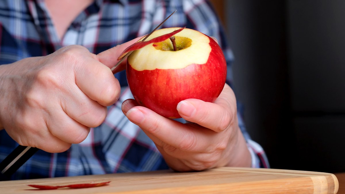 Haende schaelen einen roten Apfel mit einem kleinen Messer ueber einem Holzbrett