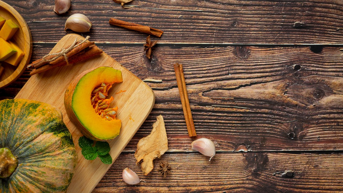Aufgeschnittener Kuerbis auf einem Holzbrett, daneben ein ganzer Kuerbis, Knoblauch und Zimtstangen