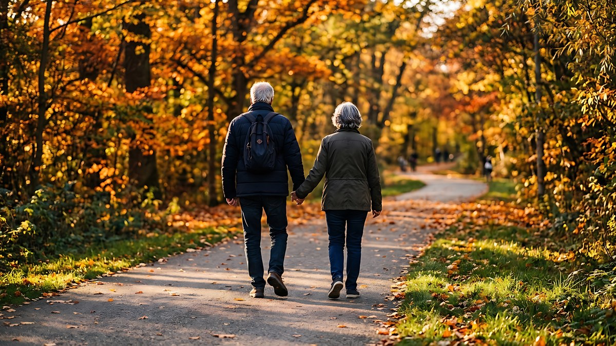Aelteres Paar geht Hand in Hand auf einem Parkweg zwischen herbstlich gefaerbten Baeumen