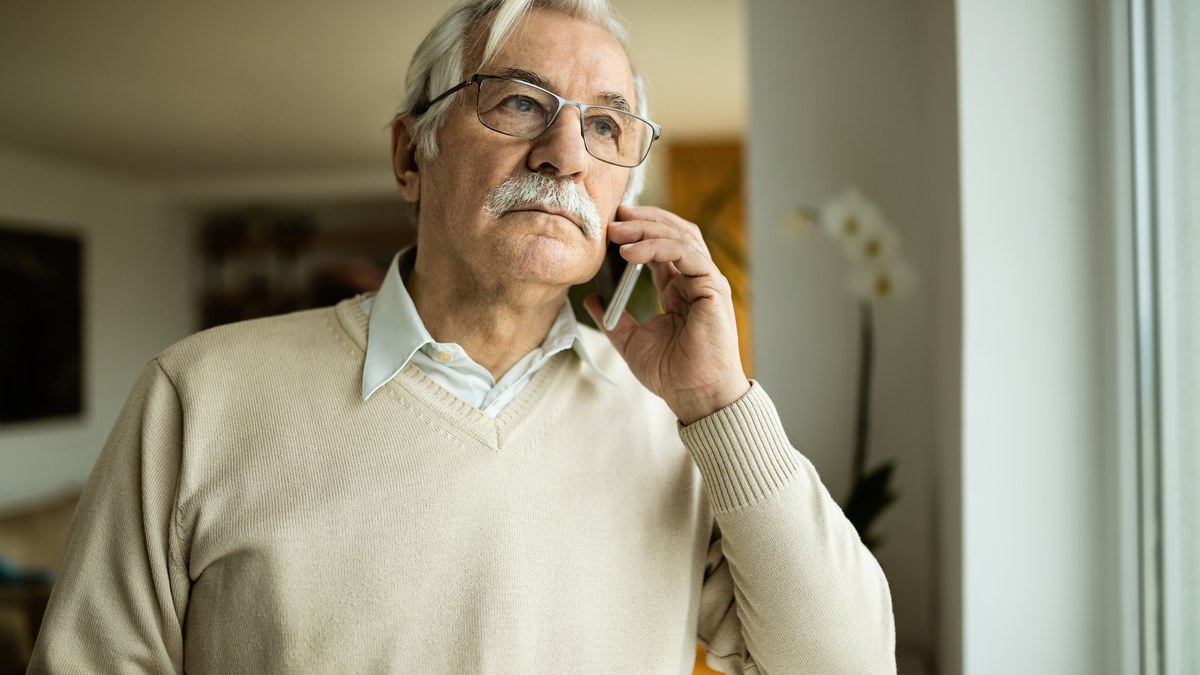 Älterer Mann mit Brille steht am Fenster und telefoniert ruhig mit dem Handy