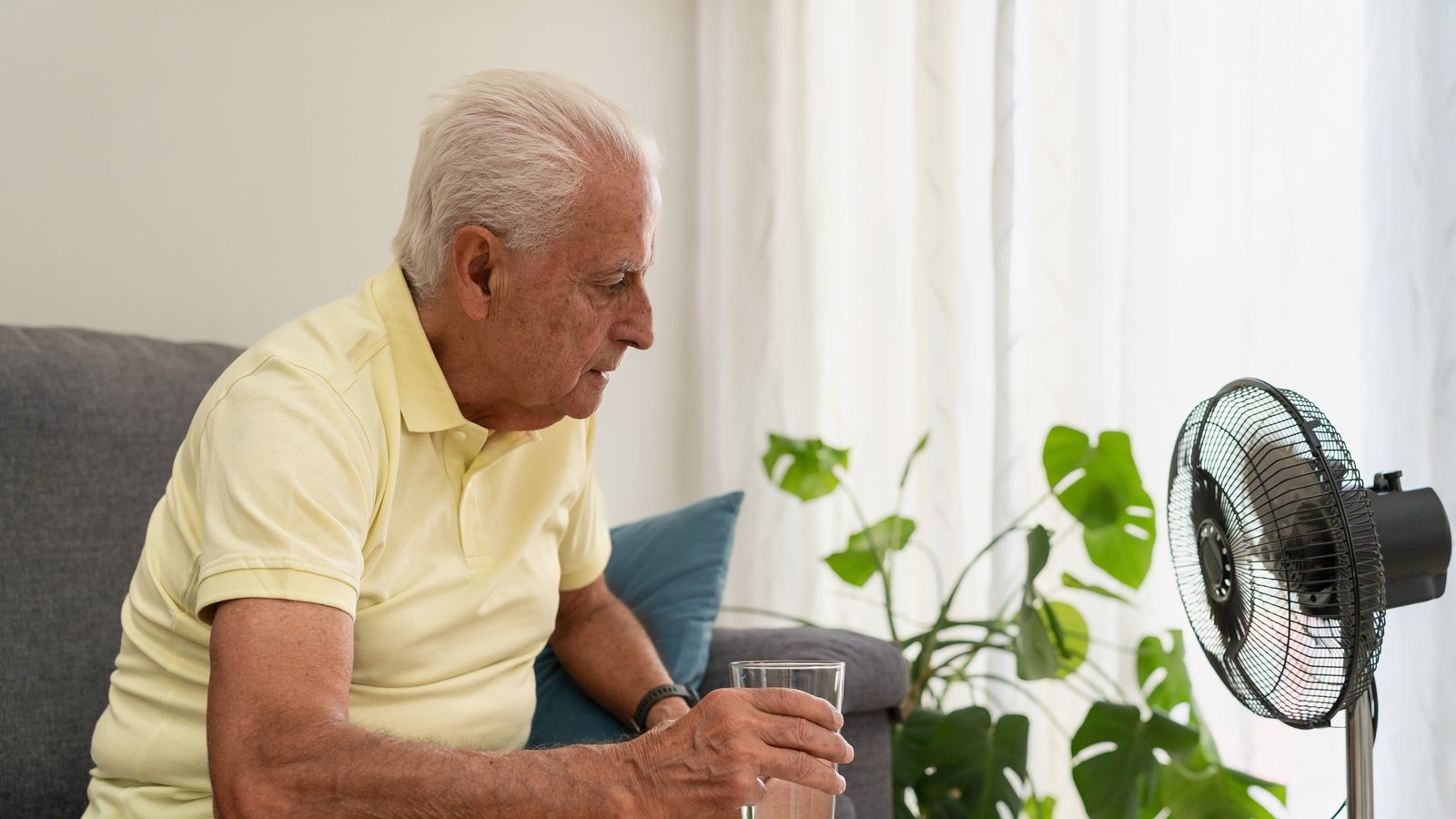 Aelterer Mann sitzt an einem heissen Tag vor dem Ventilator auf dem Sofa und haelt ein Glas Wasser