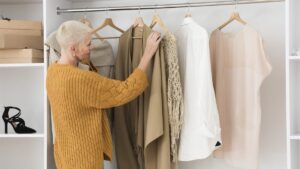 Aeltere Frau am offenen Kleiderschrank greift nach einer Strickjacke, daneben haengen Bluse und Kleid