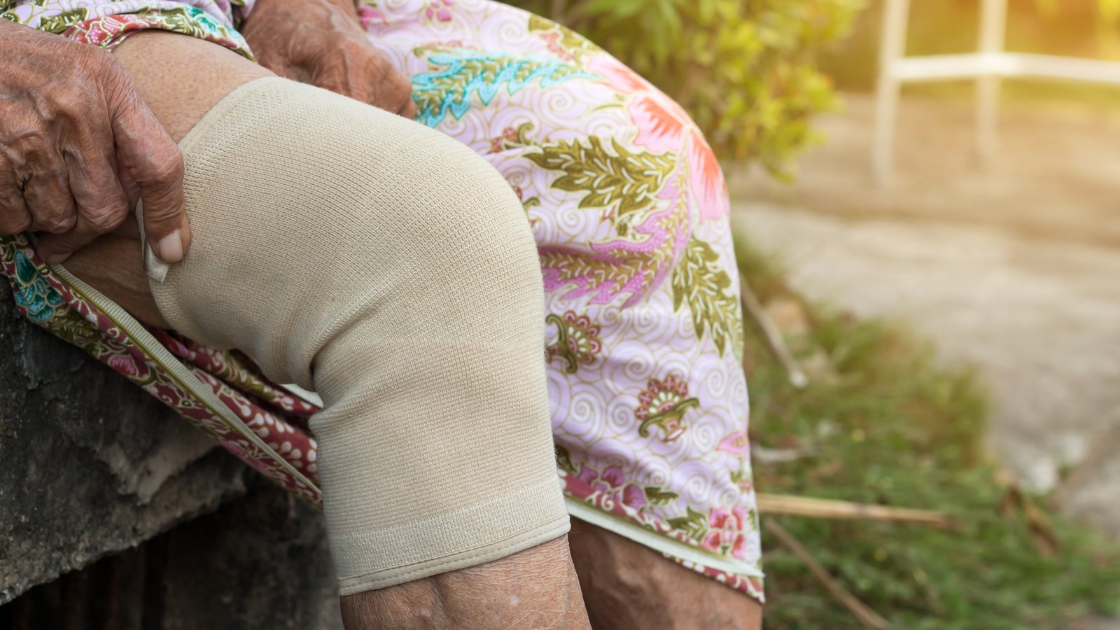 Aeltere Frau sitzt draussen und traegt eine hautfarbene Kniebandage ueber dem Knie