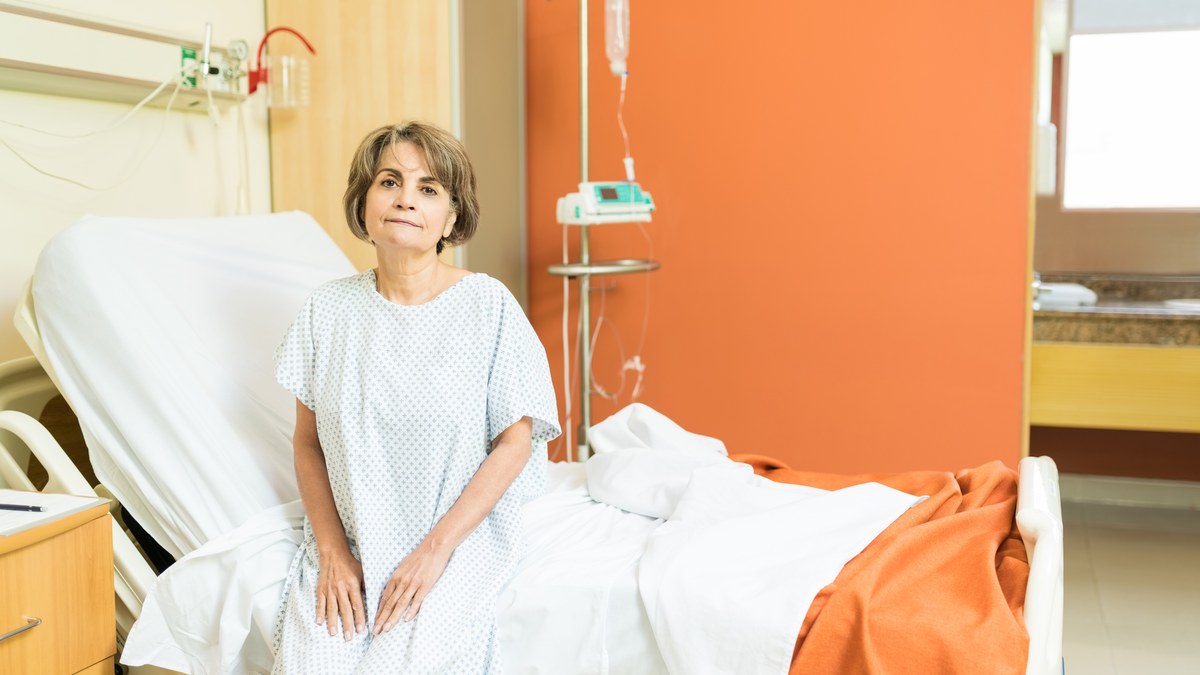 Seniorin im Krankenhaushemd sitzt auf dem Bettrand im Patientenzimmer
