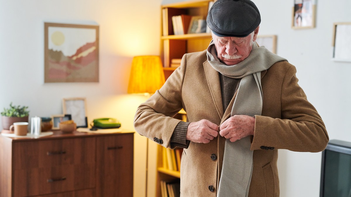 Aelterer Herr mit Schiebermuetze und Schal knoepft zu Hause seinen Wintermantel zu