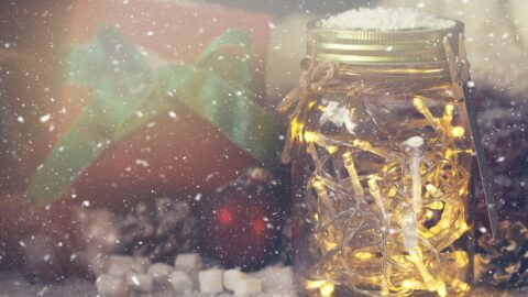 Ein Glas mit einer warmweiß leuchtenden Lichterkette neben einem Geschenk, davor fällt Schnee