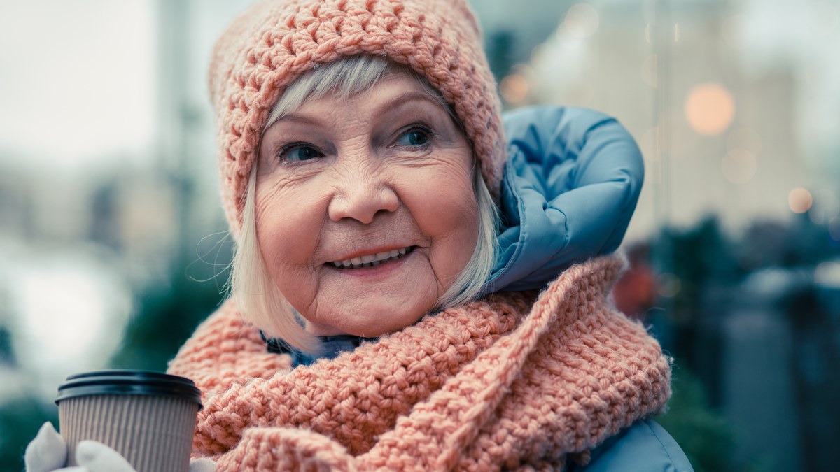 Aeltere Dame mit grober Strickmuetze und passendem Schal haelt einen Becher Kaffee und laechelt