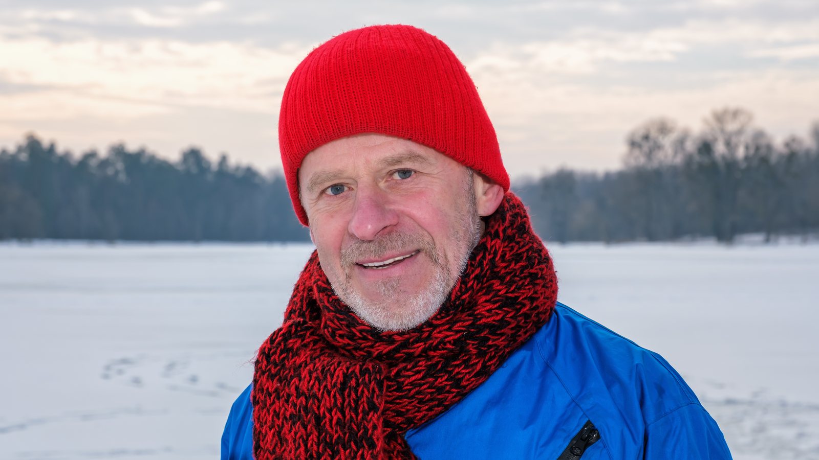 Aelterer Mann mit roter Strickmuetze und Schal steht laechelnd in einer verschneiten Landschaft