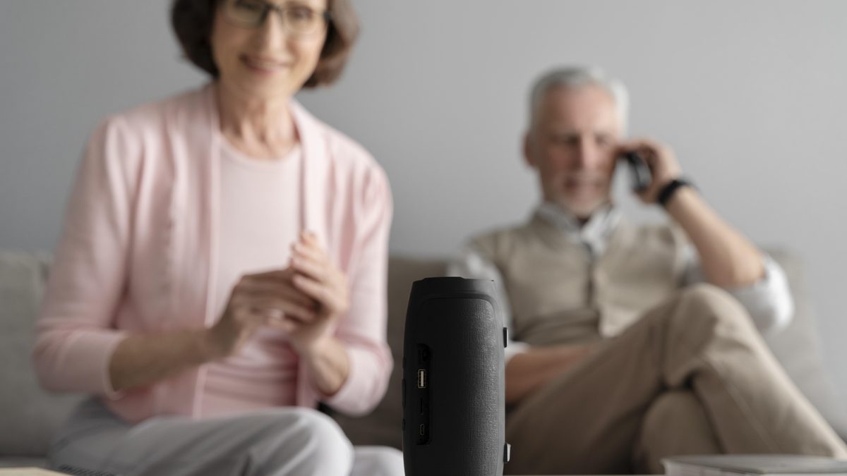 Aeltere Frau sitzt laechelnd im Wohnzimmer, vor ihr steht ein kleiner Lautsprecher auf dem Tisch