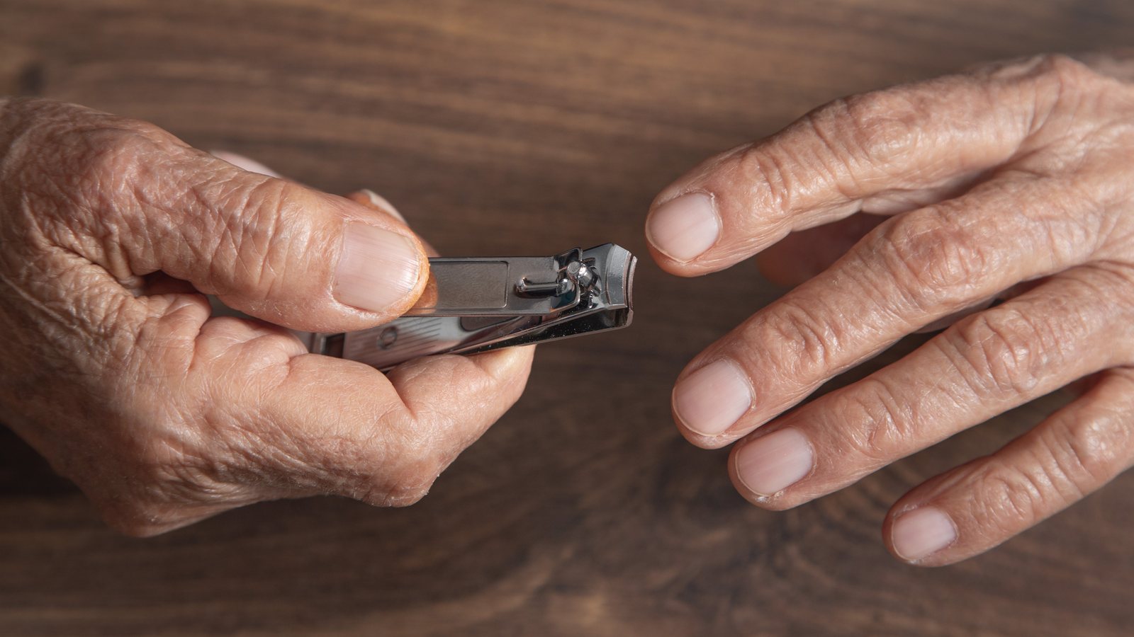 Aeltere Haende halten einen Nagelknipser aus Metall und kuerzen ueber einem Holztisch die Fingernaegel