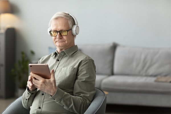 Älterer Mann mit Kopfhörern und Tablet im Sessel im Wohnzimmer