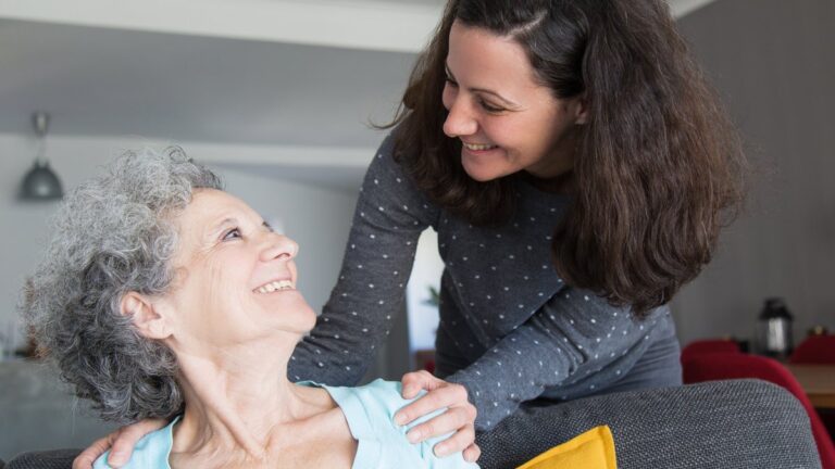 Tochter besucht ihre aeltere Mutter und legt ihr die Haende auf die Schultern, beide laecheln