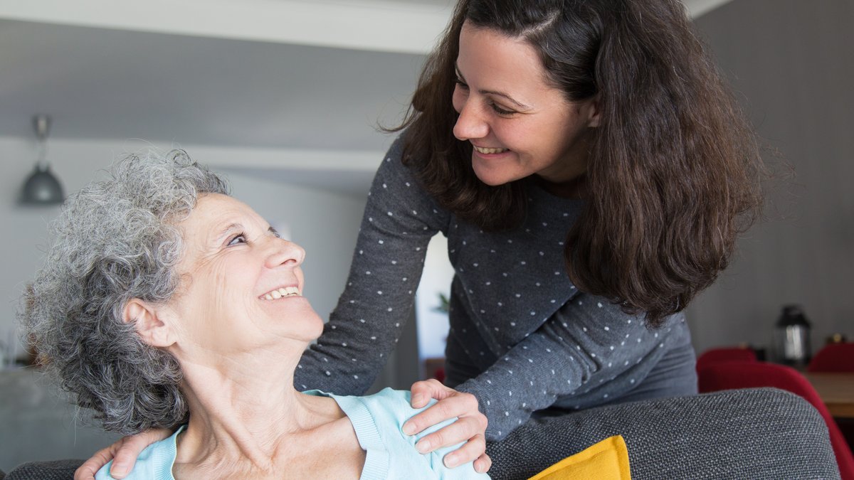 Tochter besucht ihre aeltere Mutter und legt ihr die Haende auf die Schultern, beide laecheln