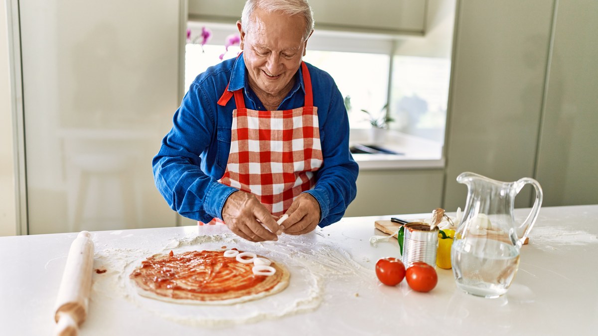 Aelterer Mann mit Schuerze belegt in seiner Kueche laechelnd eine Pizza