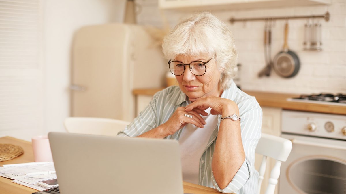 Aeltere Frau mit Brille sitzt in der Kueche vor einem Laptop und liest aufmerksam