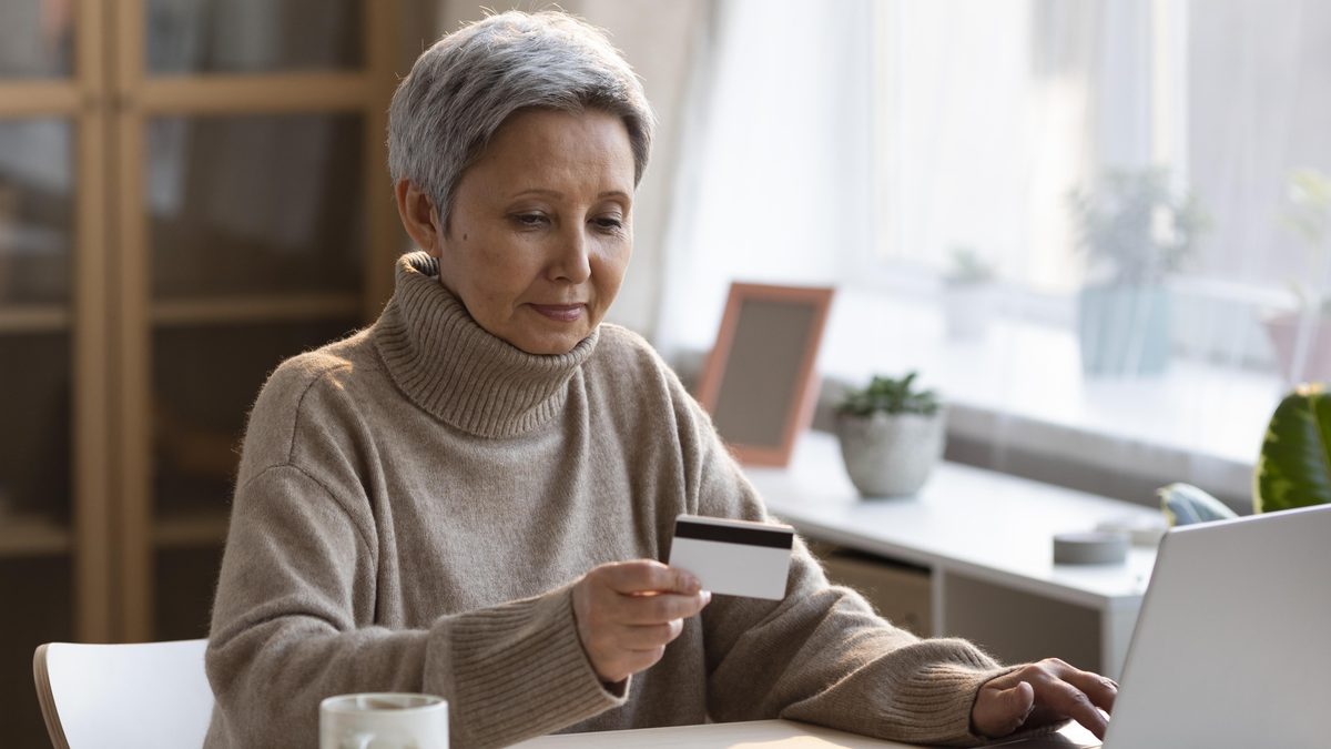 Aeltere Frau sitzt am Tisch vor einem Laptop und haelt eine Bankkarte in der Hand