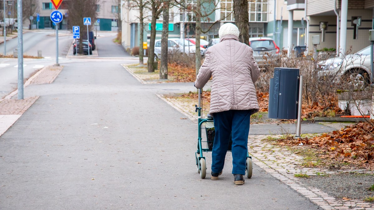 Aeltere Frau mit Rollator geht an einem grauen Herbsttag auf dem Gehweg