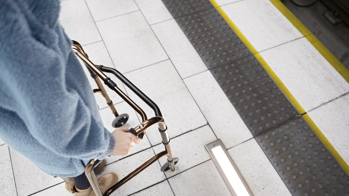 Haende an einer Gehhilfe mit Raedern vor einer Bahnsteigkante mit gelber Markierung