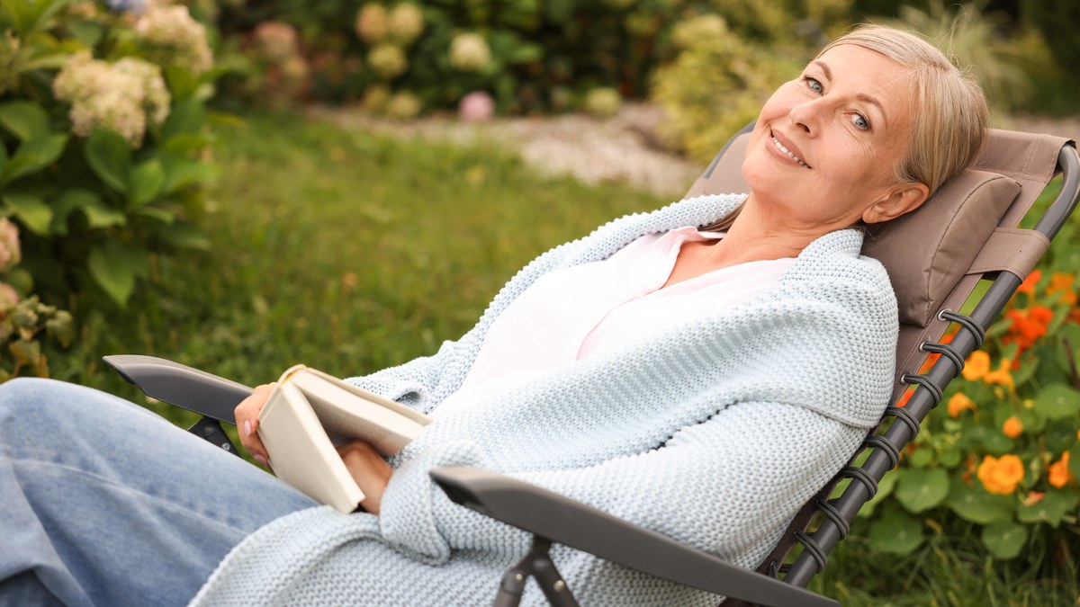 Aeltere Frau mit Buch und Strickjacke liegt entspannt in einem Gartensessel zwischen Hortensien
