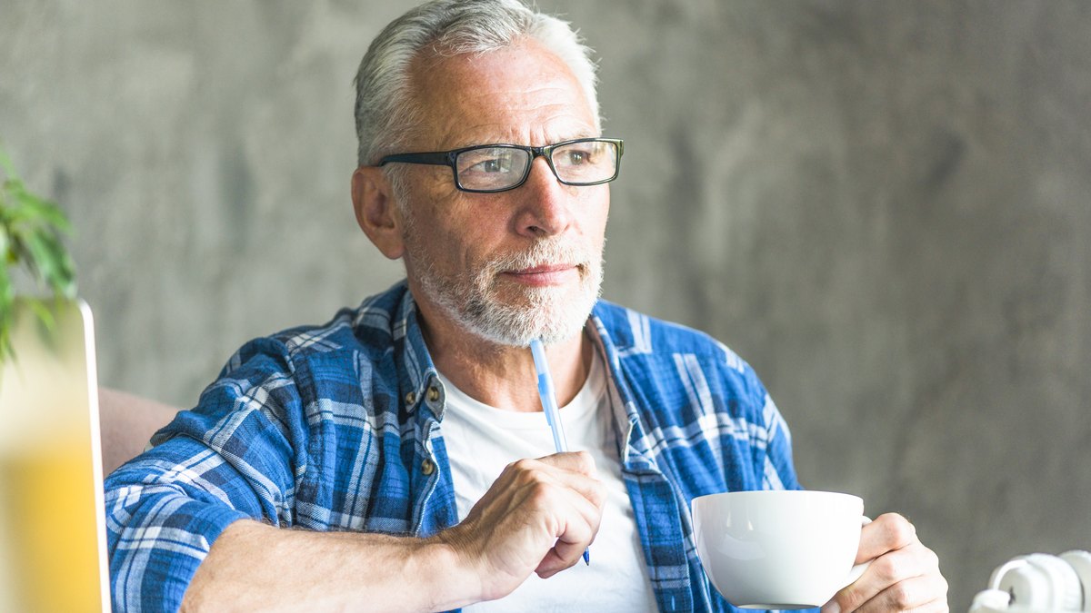 Aelterer Mann mit Brille und Stift am Tisch, denkt nach, bevor er etwas aufschreibt